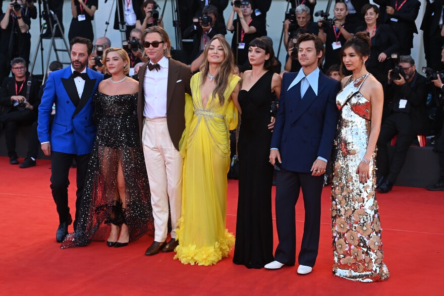 A row of actors have their arms around each other while in formal dress on a red carpet. Paparazzi is in the background.