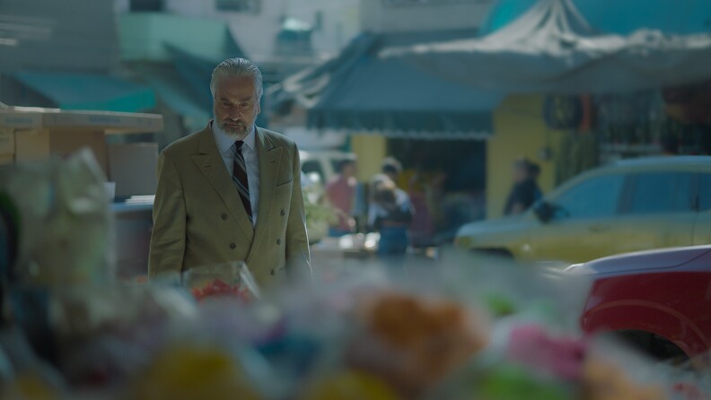 A image from a film still of an older man with white hair wearing a double-breasted tan suit, white shirt and tie. He is standing outside on a crowded street.