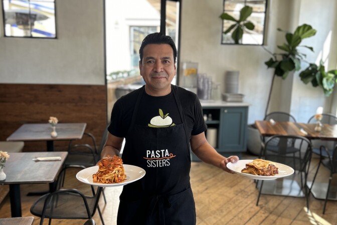 A man with a medium-dark skin tone, black hair, a black T-shirt, and an apron that reads "Pasta Sisters" in embroidered white lettering stands in the middle of a restaurant. He's holding two plates of food. One plate contains long noodles dressed in a red sauce piled high in a small tower-like formation. The other plate contains flat pasta noodles with red meat sauce, resembling lasagna. In the background are various wood-topped tables and chairs, some containing place settings and small vases of flowers.