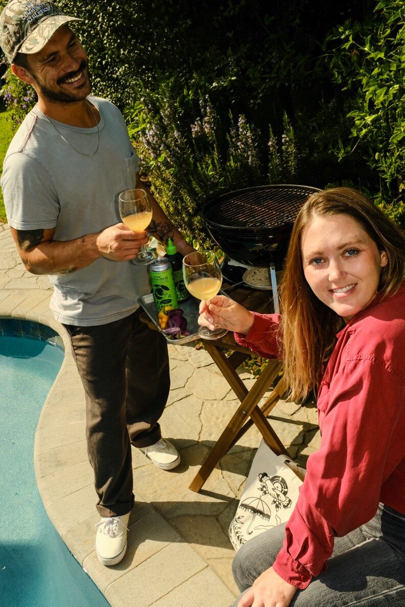 A man with medium dark skin in a cap and t-shirt and a woman with light skin in a red shirt share drinks next to a backyard grill by the pool.