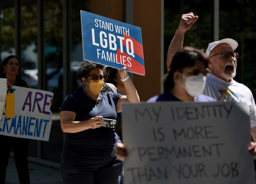 People march holding signs in support of LGBTQ+ people. The signs' messages include "MY IDENTITY IS MORE PERMANENT THAN YOUR JOB" and "STAND WITH LGBTQ FAMILIES." Several women are marching, with a couple in masks, others not. An older man in white marching with the group wears no mask and has his fist raised.