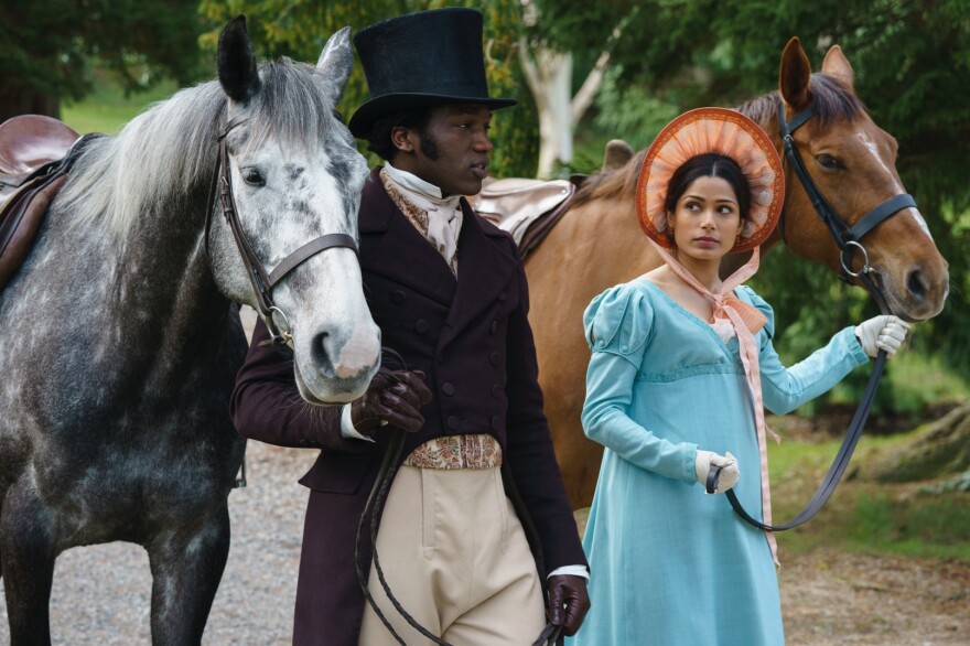 Ṣọpẹ́ Dìrísù (left) dressed in a regal Victorian era suit with sideburns and a large top hat leads a grey horse by the bridle. To his right, Freida Pinto wears a blue dress of similar styling and a orange bonnett, and she leads a brown horse. Both are walking to the right on a gravel path, shown from the knee up, with Freida looking at Ṣọpẹ́ . In the background, a slightly wooded grassy area.