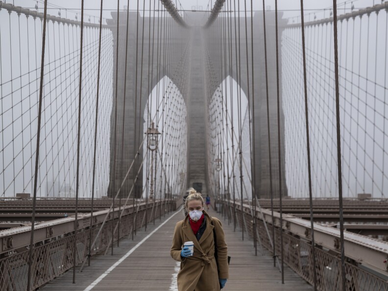 A masked person stands along on a bridge holding a coffee cup.