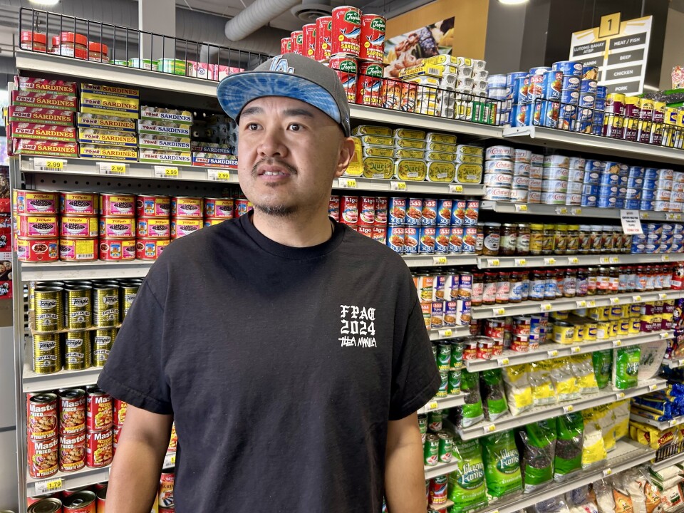 A male presenting person wears a baseball cap. He stands in front of super market shelves with canned goods.