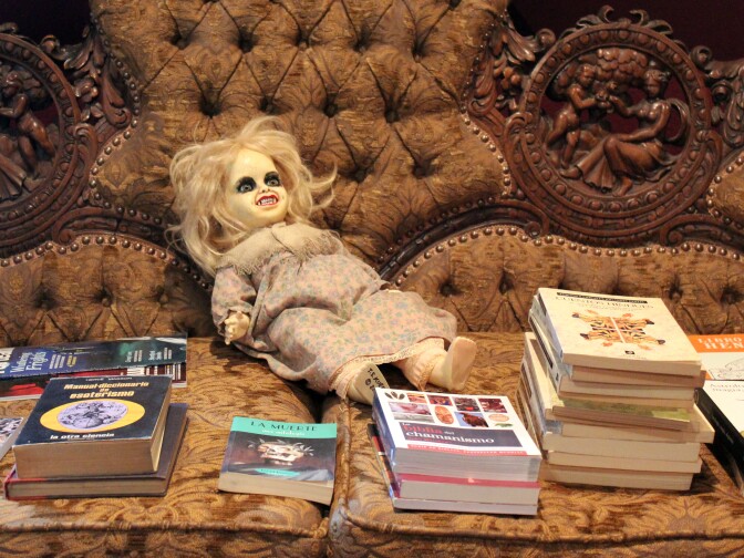 A couch piled with books and a demonic doll. 