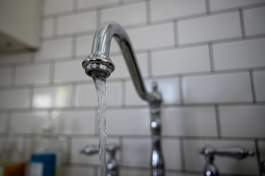 Water streams from a faucet. White brick-shaped tiles with dark grout line the wall behind it.