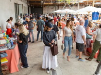 Several dozen people walk across a courtyard buying books. A woman in the foreground wears a blue hat, blue sweatshirt, a white skirt, and carries a brown bag. She is putting something into the bag. People can be seen walking and in conversation behind her.