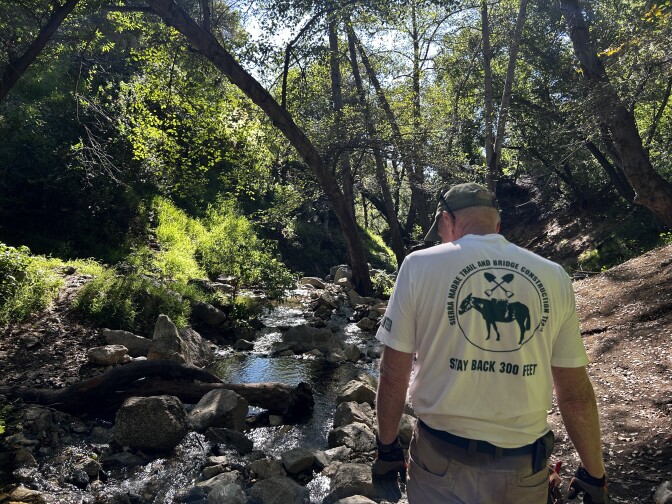 The back of an older white man wearing a white shirt that reads "sierra madre trail and bridge construction team. stay back 300 feet" with the image of a burrow and two crossed shovels. In the background is a creek and grove shaded by trees. 