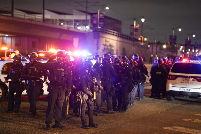 LAPD Officers line up in preparation to form a skirmish line in front of protesters near the federal detention facility in downtown L.A. on June 7, 2025. Most of the officers hold batons. One officer in the front holds a less-lethal projectile launcher.