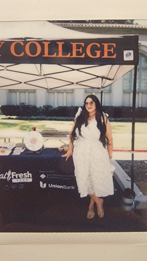 A woman with brown skin tone wearing a white dress stands at a recruiting table outside, smiling.