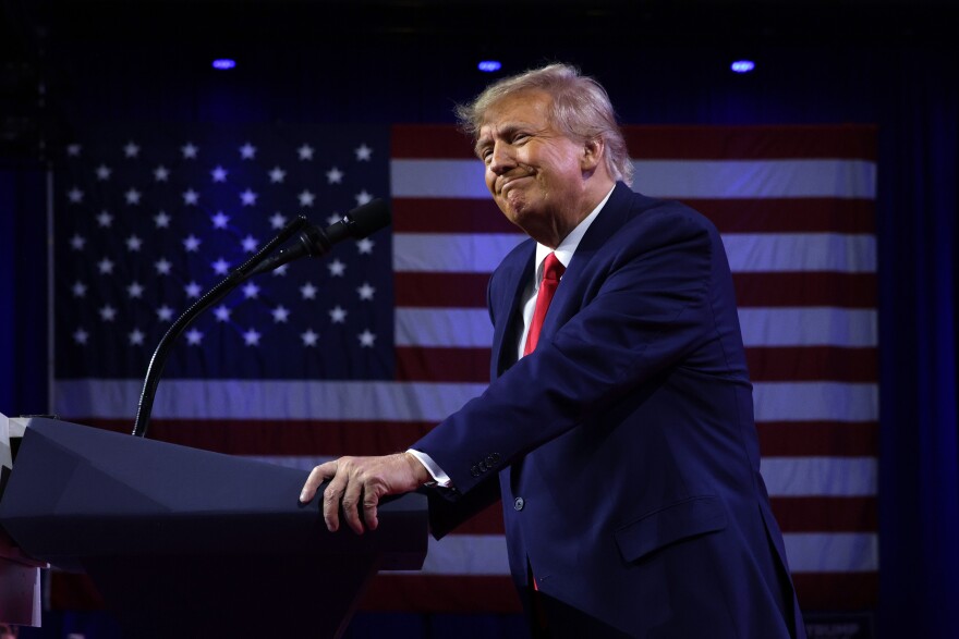 A man with light-tone skin wears in a dark suit and red tie as he leans on a lectern with a U.S. flag behind him