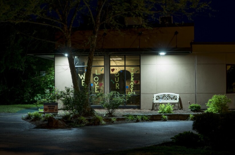 A nighttime shows the window of a nursing home with the words: WE LOVE YOUR THERAPY written vertical on each pain. An empty bench is to the right of the windows.
