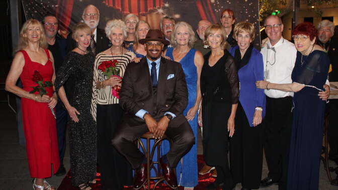 A group in formal attire poses together at an event, with one person seated on a stool at the center.