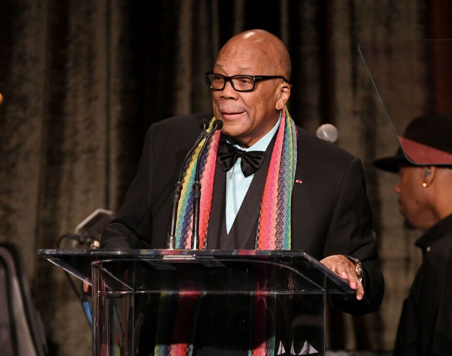 Quincy Jones, dressed in a tux and wearing a multicolored scarf, speaking at a podium.