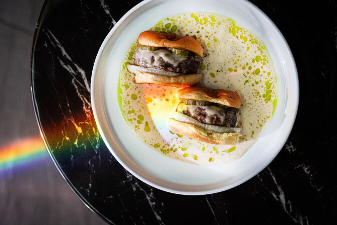 A dry-aged beef burger sliced in half, served in a light broth on a white plate with a rainbow light reflection across a dark marble table.