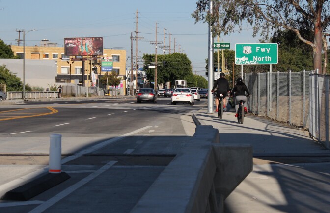 Two people ride bikes on a sidewalk as people drive cars on the road next to them.