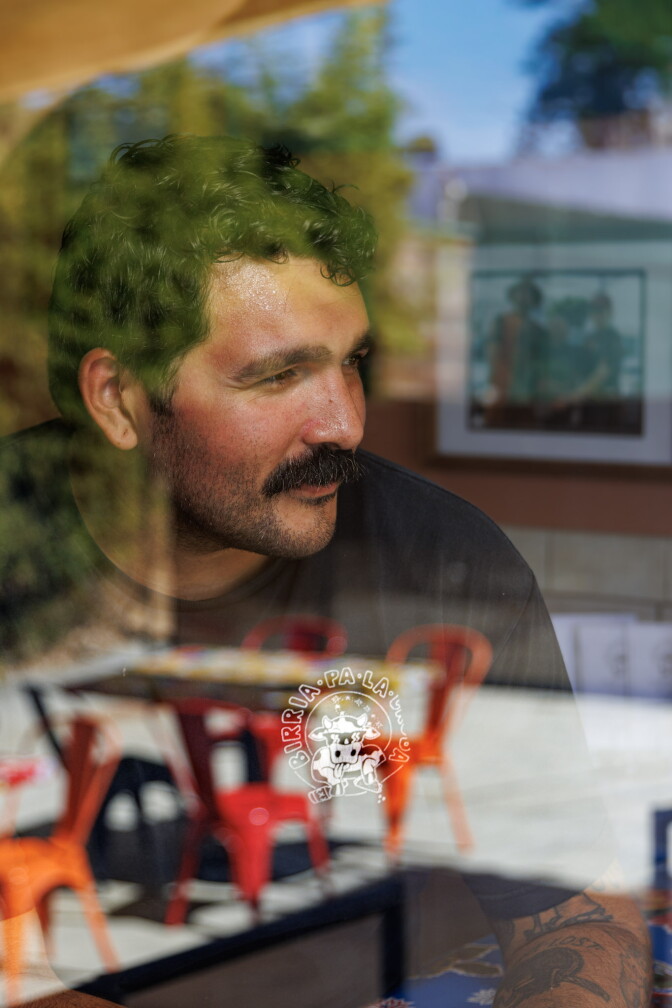 A man with a medium-dark skin tone, curly dark hair and a mustache looks out through a window bearing the Birria Pa La Cruda logo, a cartoon cow. Red and orange metal chairs and trees are reflected in the glass.