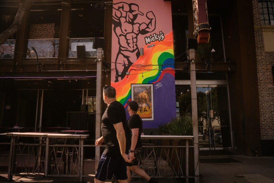Two people with light skin tones walk down a sidewalk looking away from the camera. Next to them is art for Micky's, a gay club. It has an illutration of a muscular man and wavy rainbow pride colors.
