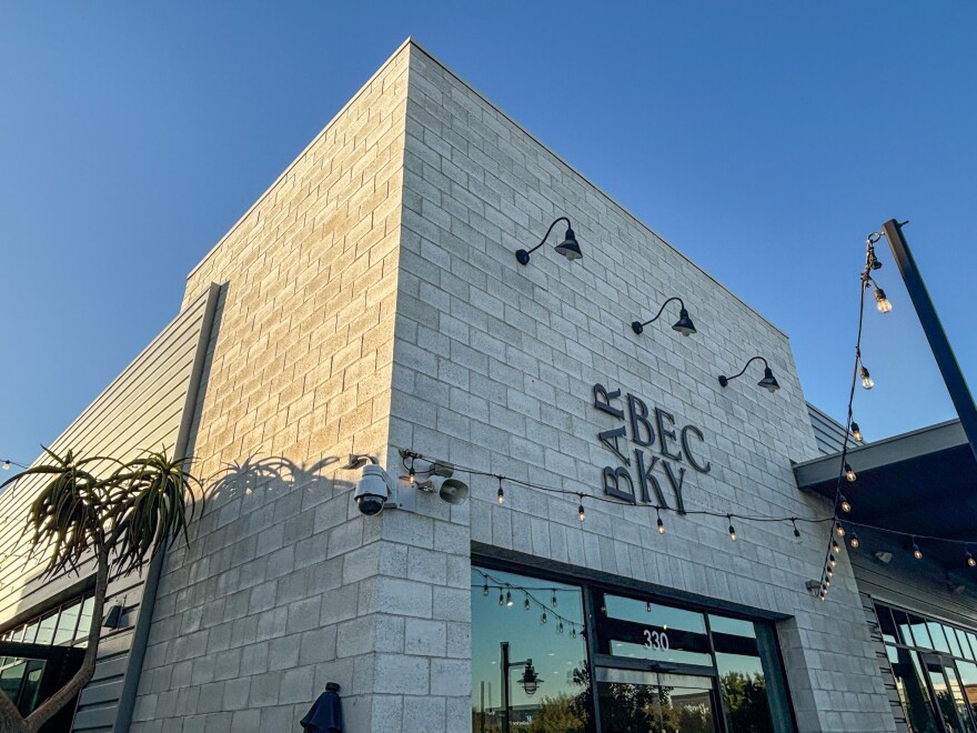 The exterior of Bar Becky, a modern gray brick building with black lamps, string lights, and a clear blue sky above.