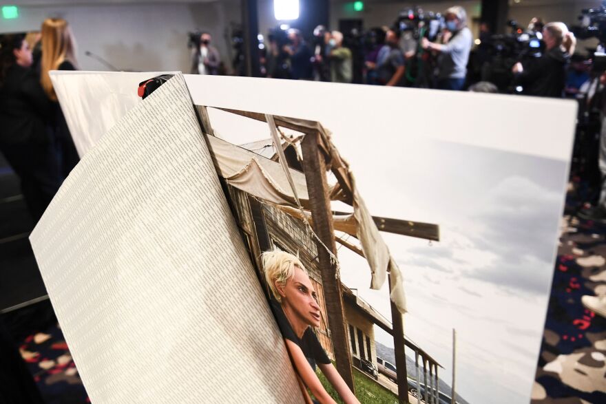 An enlarged photo of a woman with short blonde hair sitting on a rustic porch is in the foreground with a news conference taking place in the background.