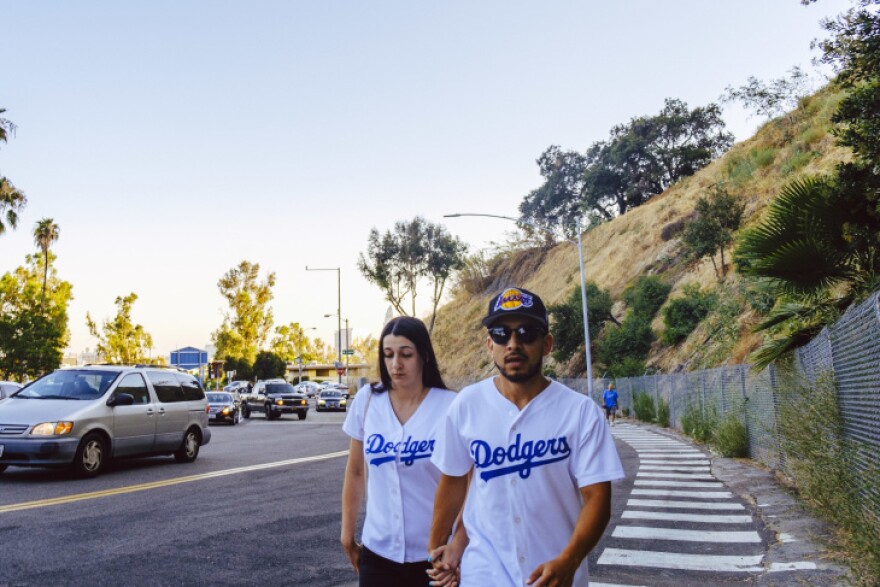 A man and woman in white Dodgers jerseys hold hands as they walk up the pedestrian pathway to the ballpark. One wears a Lakers cap. To the far left: Lines of cars also waiting to get into the ballpark.