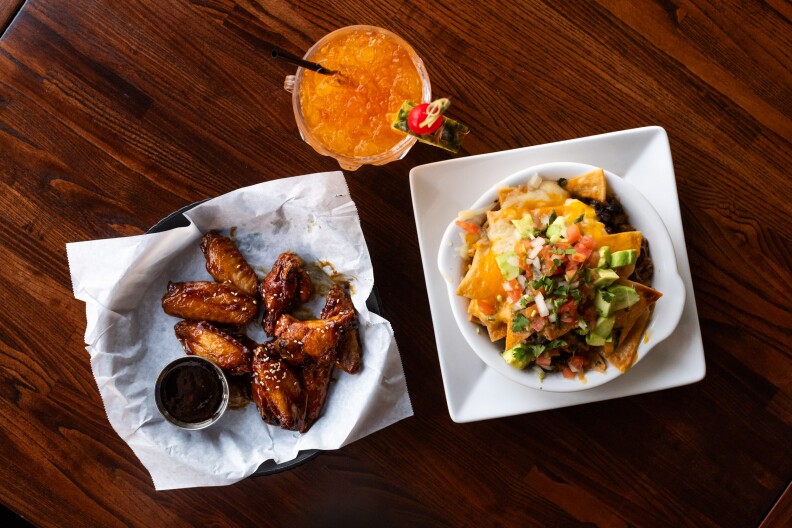 An overhead photo of a plate of dark orange wings, a white square plate with nachos, and an orange cocktail.