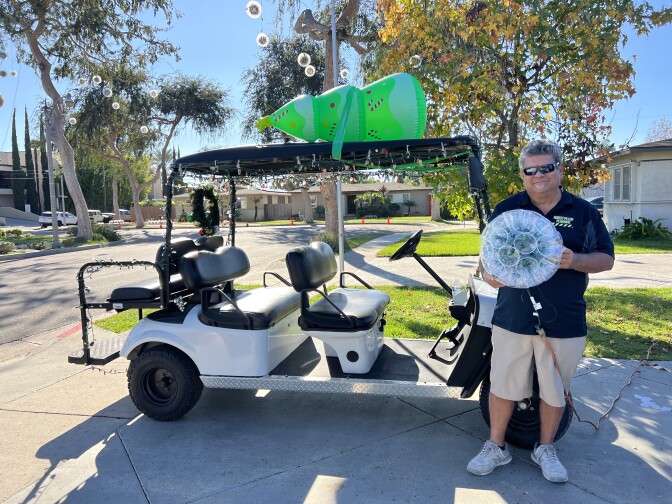 A man wearing khaki shorts, a black tshirt and sunglasses holds up a sparkle ball in front of a golf cart. 