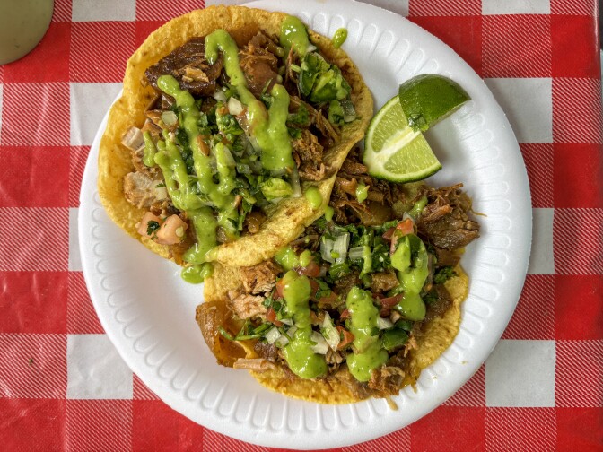Two corn tortilla tacos on paper plate filled with carnitas, topped with bright green salsa, cilantro, onions, and chicharrón, with lime wedges on side