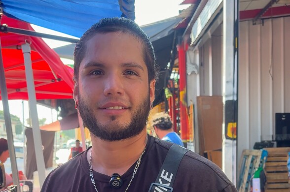 A medium skinned man with a beard wearing a black shirt, a silver necklace, and a fanny pack with a sign that reads “NIKE.” He has tattoos on both arms.