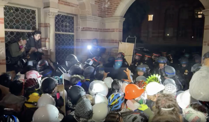 Police in riot gear push back protesters in hard hats and helmets.