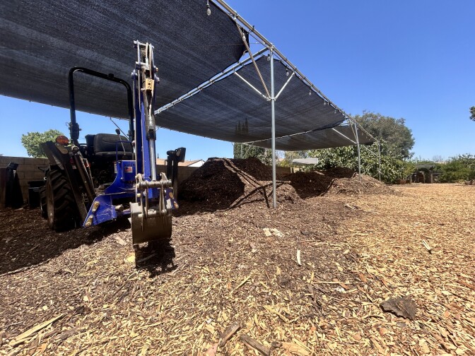 A blue and black piece of farming equipment is sitting idly under a shaded structure on a sunny, clear morning. Several foot-high mounds of mulch are scattered about next to it, and the ground is covered in a thick layer of the wood-like material.