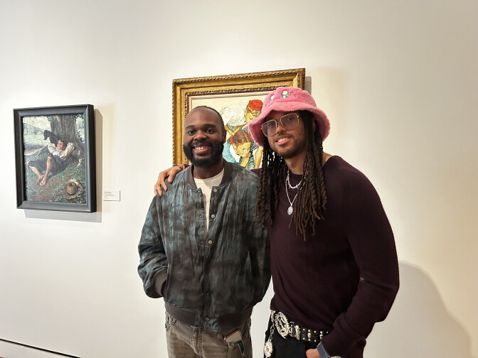 Two men smile for a photo in an art gallery, standing beside framed paintings on a white wall.