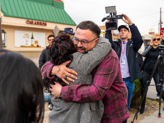 Young man in pink shirt and glasses hugs a woman in a blazer, while people take photographs behind them. 