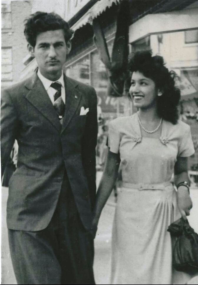 A black-and-white photo from the 1940s: a medium-skinned, dark-haired man in a suit and tie is holding hands with a medium-skinned woman with dark hair wearing a dress and pearls.