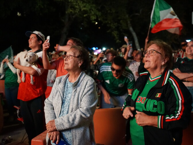 Two medium-light skin toned women wearing glasses stare at a large outdoor screen in anticipation. One of them wears a Mexico zip-up sweatshirt and Mexico shirt. Behind them, someone waves a large Mexico flag.