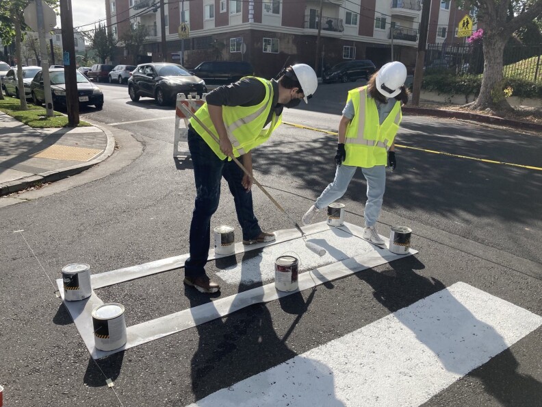 Two people wearing helmets, reflective vests and masks work to paint a crosswalk on a street. One is holding the paint roller and the other is holding the stencil in place.