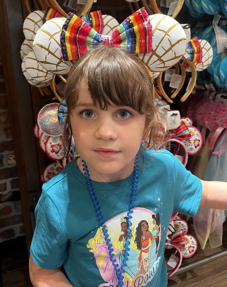 A young girl with brown hair and blue eyes looks at the camera. She is wearing Mickey Mouse ears in the shape of conchas while standing in front of a display of them. She is wearing a blue shirt with Disney characters on it. 