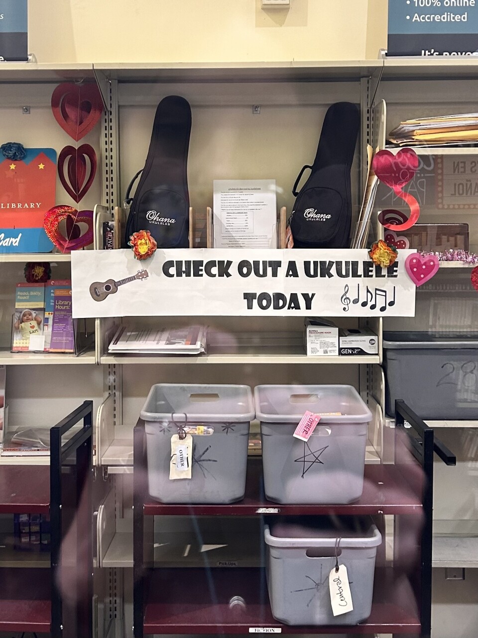 A packed wall of shelves and signs. At the center is a large white banner that says "check out a ukulele today" with two black, small cases above it. 