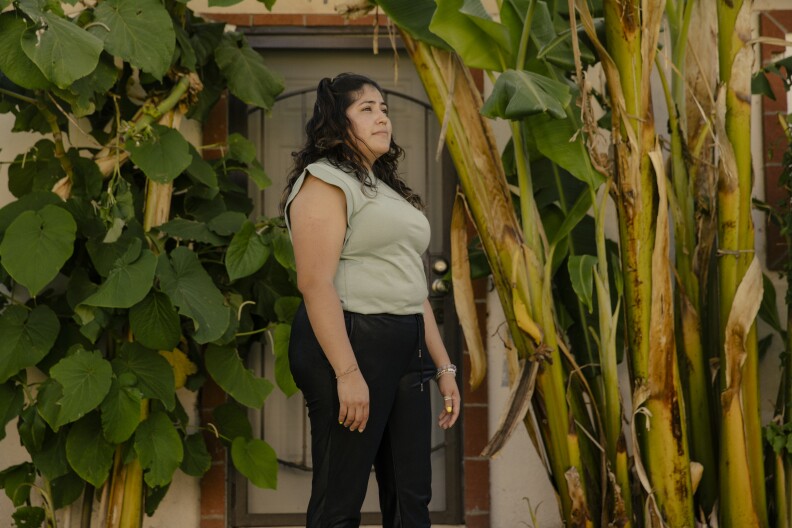 A woman with long dark hair wearing a pale green t-shirt and black pants stands in front of a doorway framed by banana plants on one side and a large-leafed plant on the other. She is looking off into the distance.