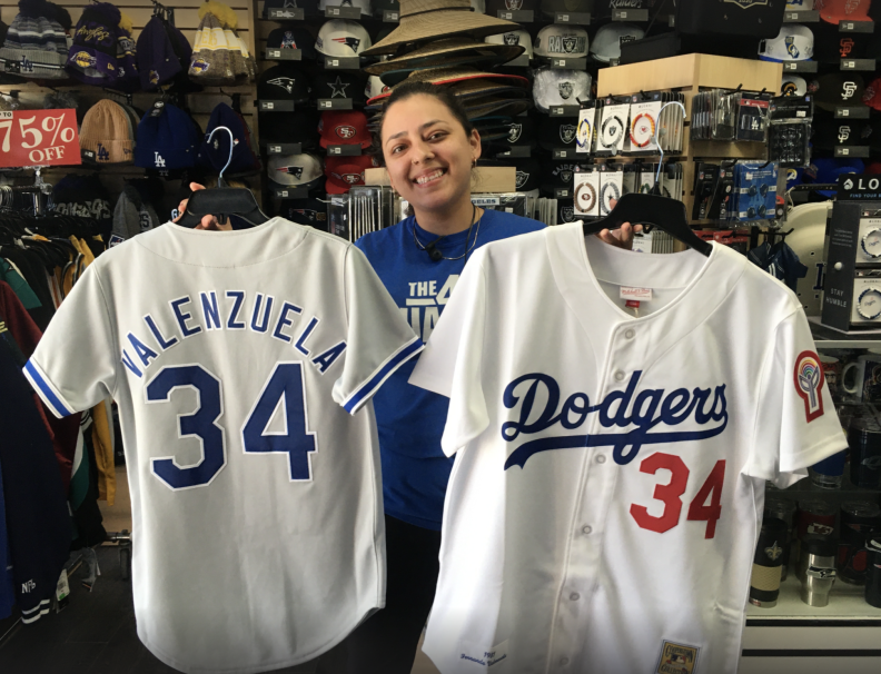 A woman smiling as she holds two white baseball jerseys, with sports hats and other sports team merchandise in the background.