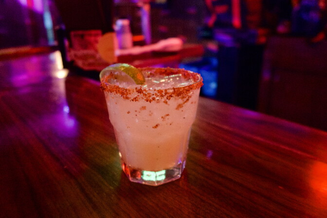 A margarita with a chili-salt rim and lime wedge sits on a bar counter lit by purple and red lights.