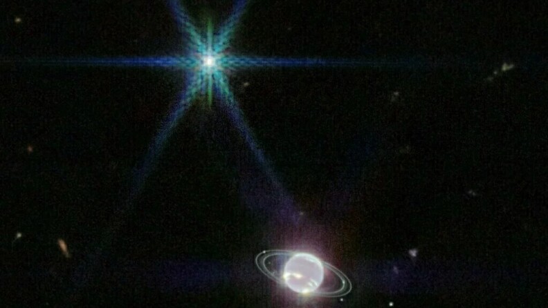 An image from outer space shows a bright star emitting light rays at the top of the photo and a small, bright white planet with two almost transparent white rings at the bottom of the photo. The background is the black sky.
