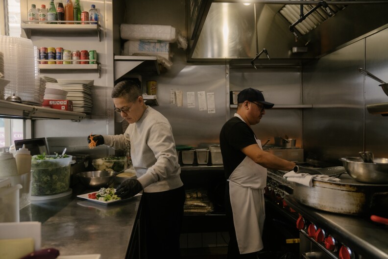 a woman with close cropped dark hair and glasses, wearing a chef's top, stands in a restaurant kitchen, carefully placing vegetables on a plate. Behind her, another man, wearing a black baseball hat and white apron, cooks on a stove