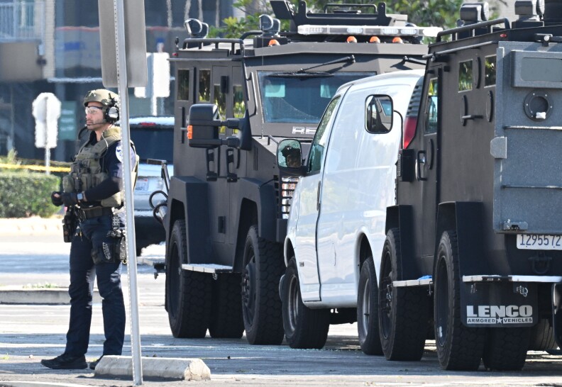 A law enforcement officer in tactical gear stands guard by a white van parker between two armored vehicles