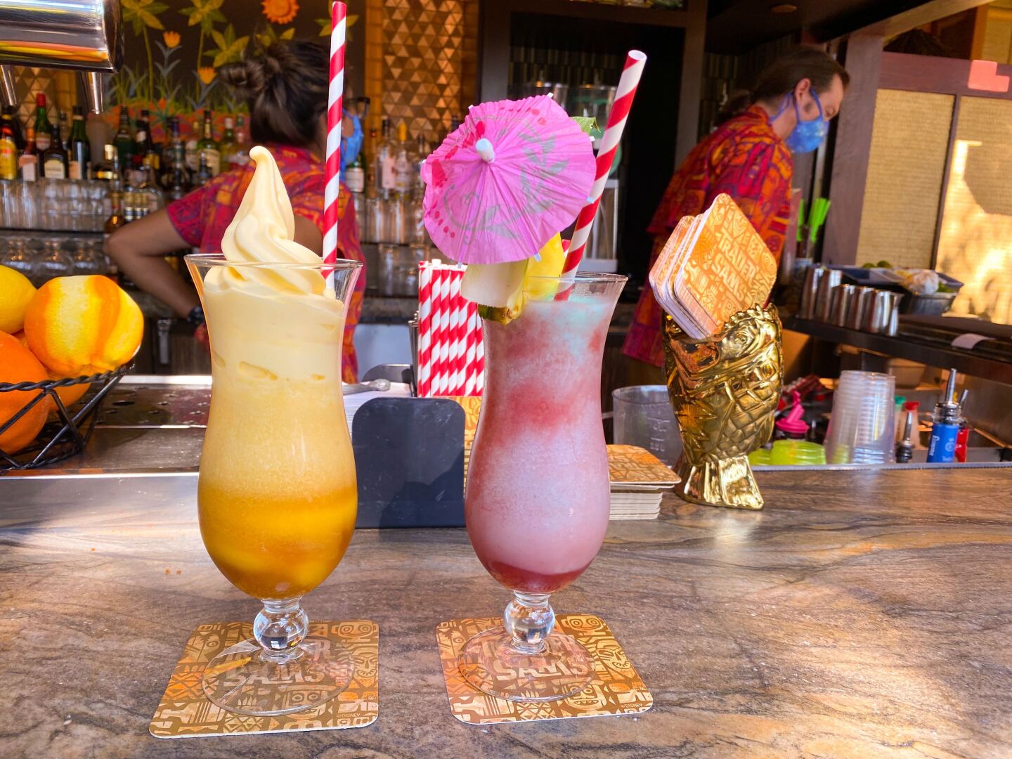 Two hurricane glasses sit on a bar. One is filled with a creamy frozen orange substance, the other is filled with a slushy pink substance.