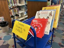 A blue cart in a library with a yellow sign that says "Relocate Juvenile to Adult." On the top shelf of the cart, you can see a red board book titled "Once Upon a Potty," and other books behind it including "The New Parent and "It's So Amazing!"