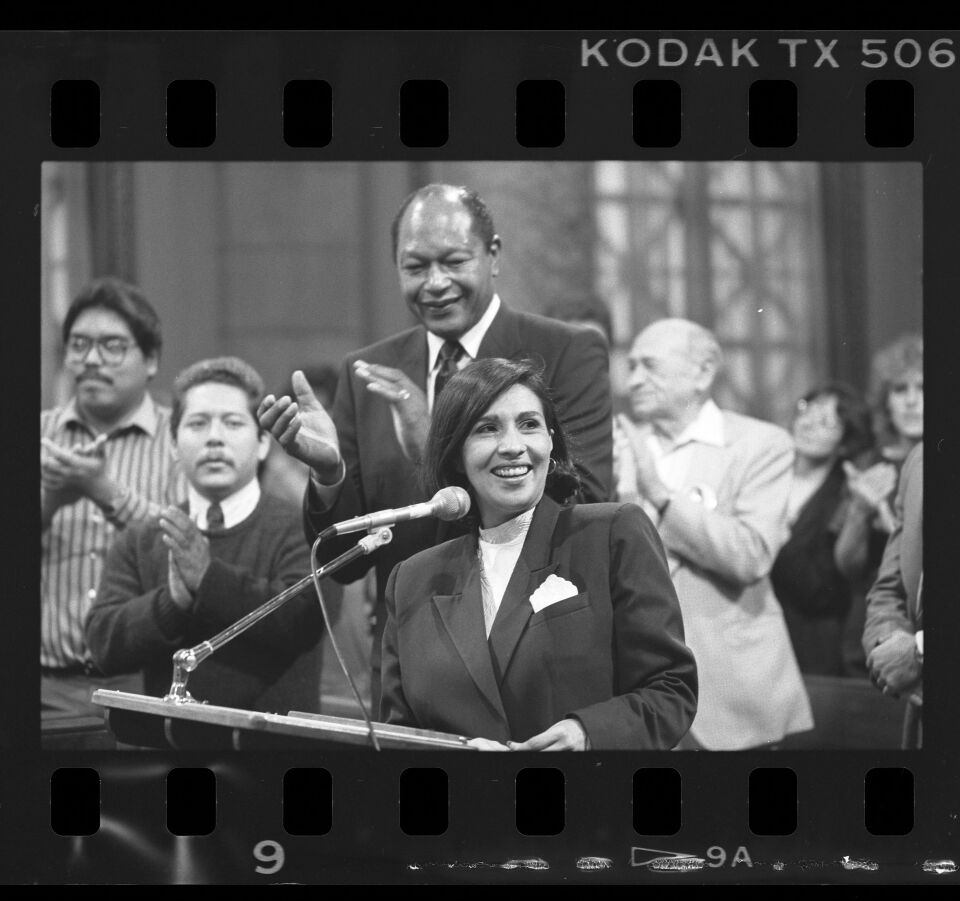 A black and white photo of a young Latina woman with short dark hair wearing a dark blazer and white blouse. She stands at a podium with a microphone and is looking towards the right of frame, smiling. Behind her there is a tall Black man wearing a suit smiling at her and clapping. Behind him there are various people also clapping. On the edges of the photo you can see the film perforations and the words "Kodak."