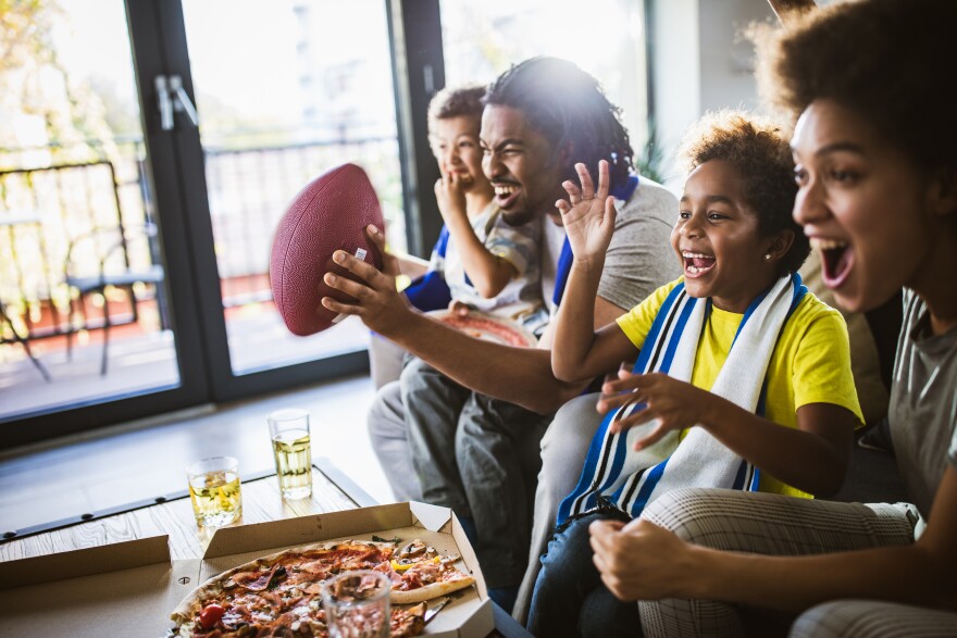 An excited Black family of two parents and two kids cheers on the couch with a box of pizza and drinks on the table in front of them.
