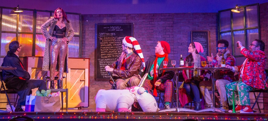A woman stands on a chair singing, while watched by six other people, some in pajamas and Santa caps
