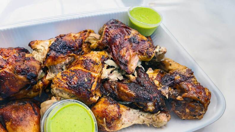 A to-go container is piled high with juicy charred chicken legs, and tucked along side it are two plastic containers filled with a dazzling herbacious green sauce for drizzling and dipping.